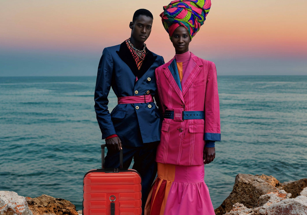 Man and woman standing on beach in bright clothes with red carry on Delsey luggage