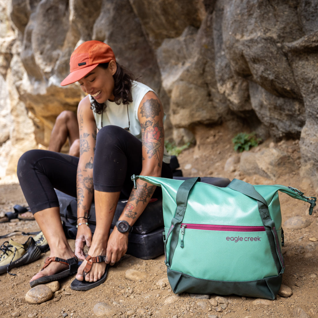 Woman sets down green Eagle Creek Migrate Tote Bag to change into sandals while hiking
