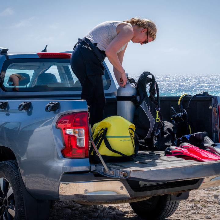 Woman unloads scuba gear from truck at beach with Eagle Creek Migrate Tote Bag in color yellow holding the snacks